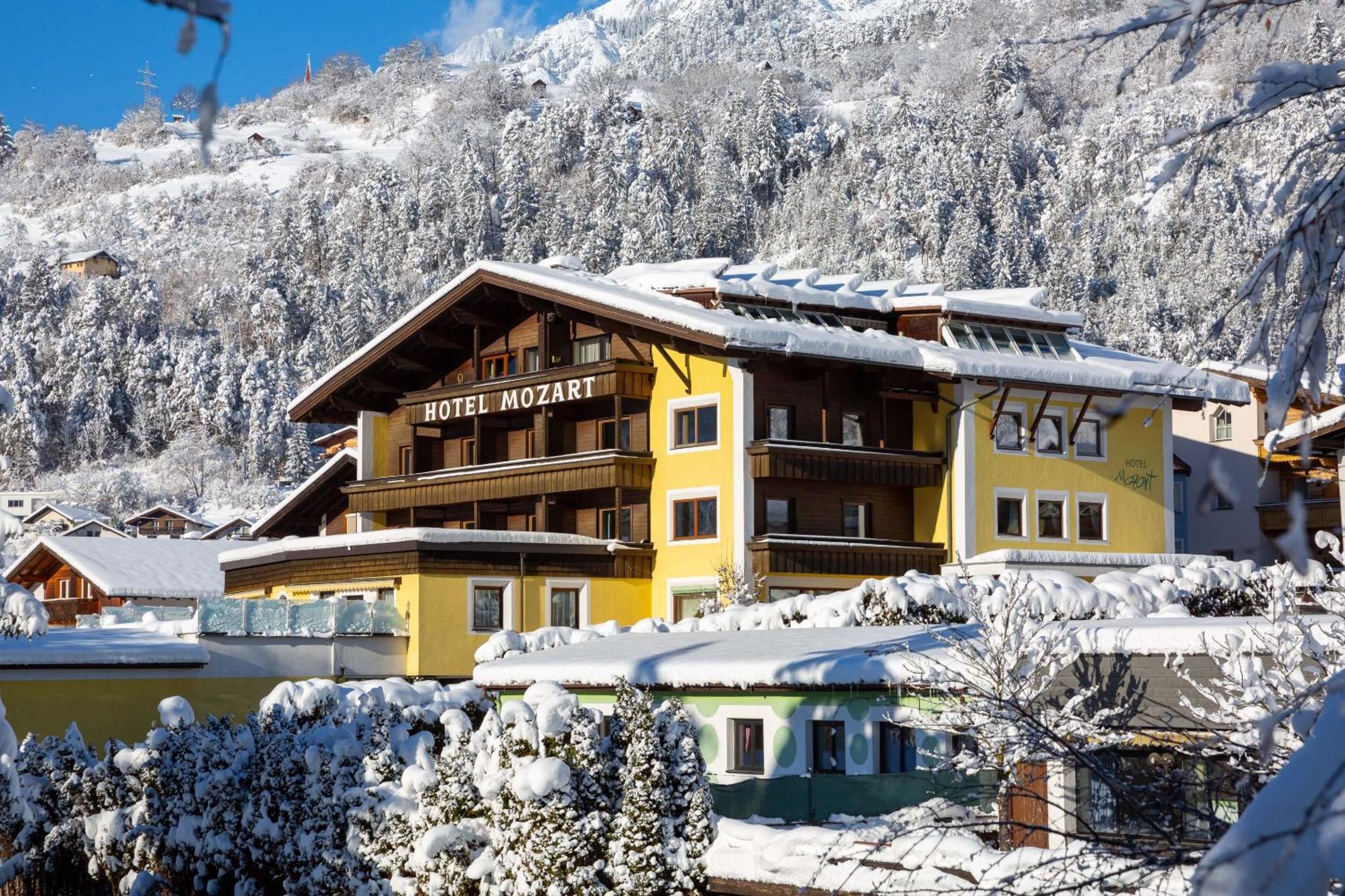 A hotel with a wooden facade and balconies, surrounded by snow-covered trees and a mountain backdrop.