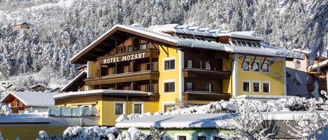 A hotel with a wooden facade and balconies, surrounded by snow-covered trees and a mountain backdrop.