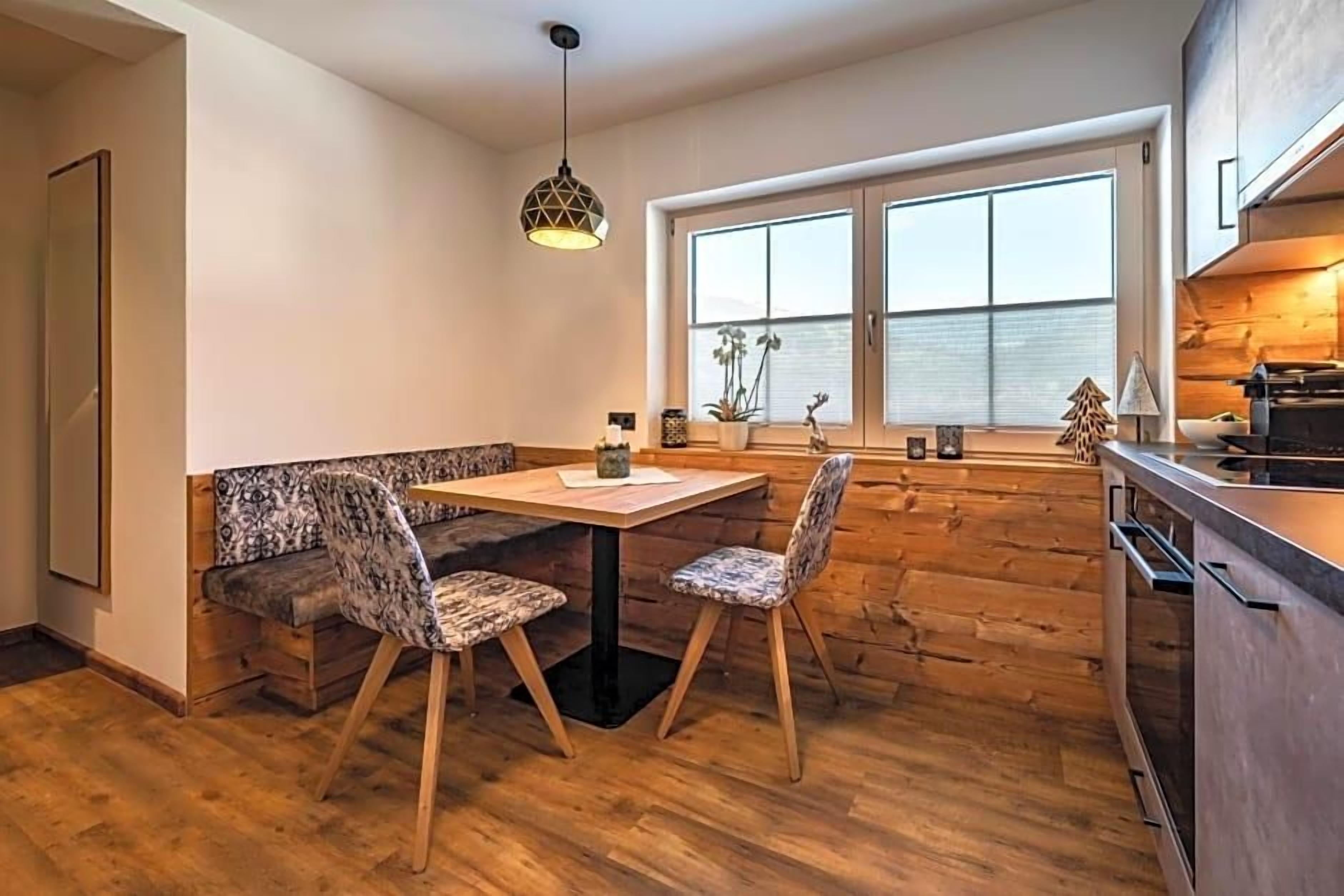 A modern kitchen with a wooden bench, two chairs, a hanging light fixture, and a window with blinds.
