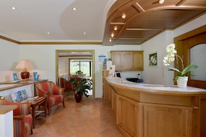 A reception area with a wooden counter, a desk with a lamp, and a chair. There is a potted plant on the counter and a window with a view of greenery outside. - Waldheim (Mayrhofen)