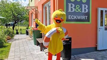 Een mascotte in een geel kostuum staat voor een gebouw met een bord waarop 'B&B ECKWIRT' staat.