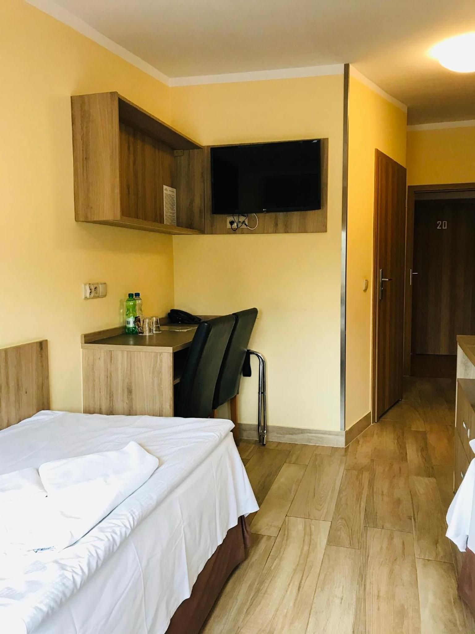 Habitación de hotel con cama, escritorio con silla, televisión montada en la pared y una puerta con número.