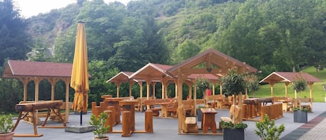 Outdoor wooden structures with benches and tables, surrounded by potted plants and a hilly landscape.