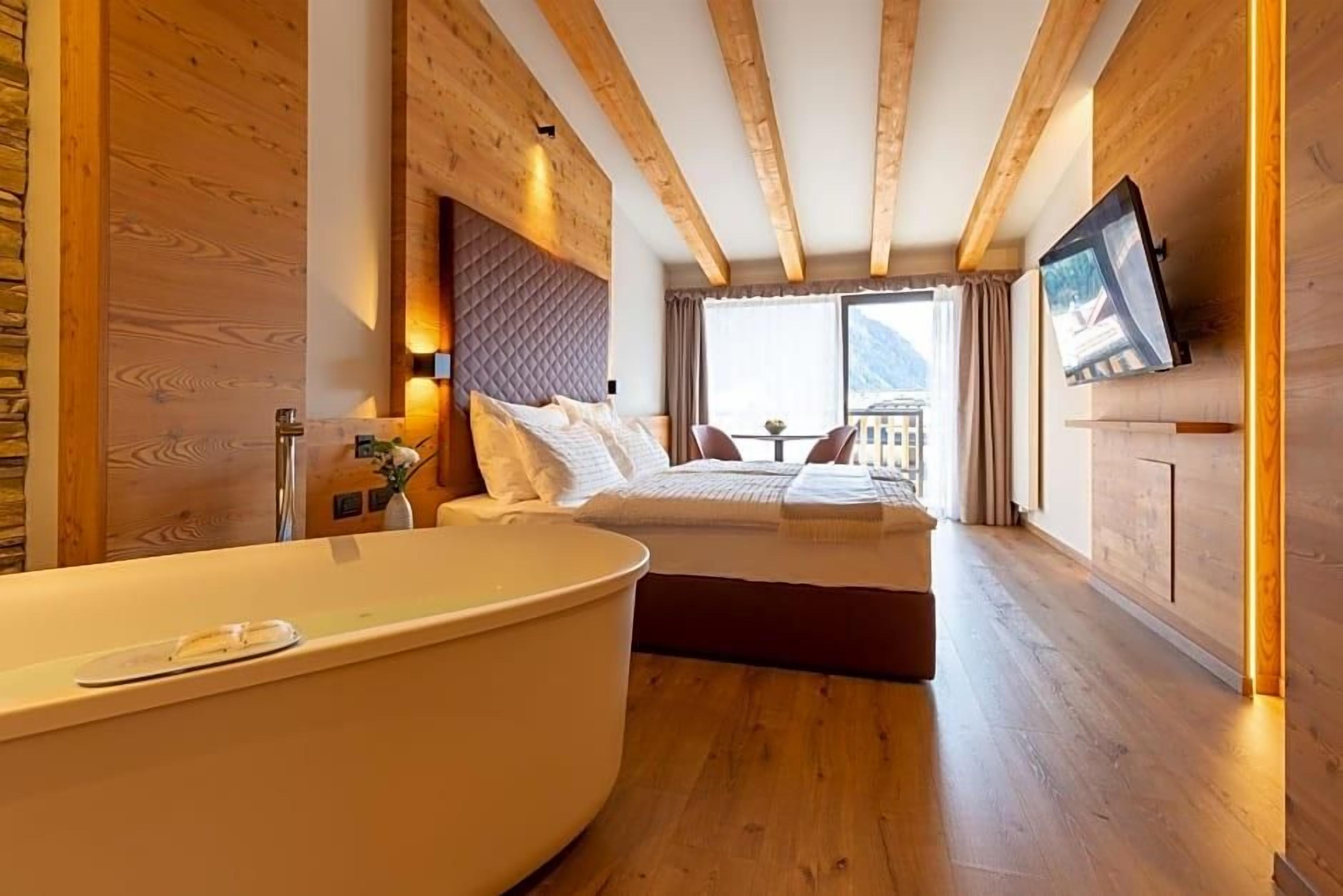 Habitació d'hotel moderna amb una banyera gran, un llit amb capçalera, una televisió penjada a la paret i un balcó amb vistes a les muntanyes.