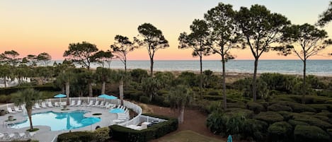 A pool area with lounge chairs and umbrellas, surrounded by well-maintained hedges and trees, overlooking a beach.