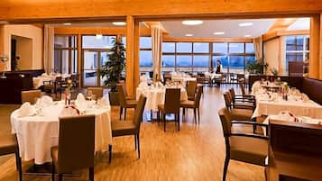 A spacious dining area with multiple tables set for guests, large windows offering a view, and wooden ceiling beams.