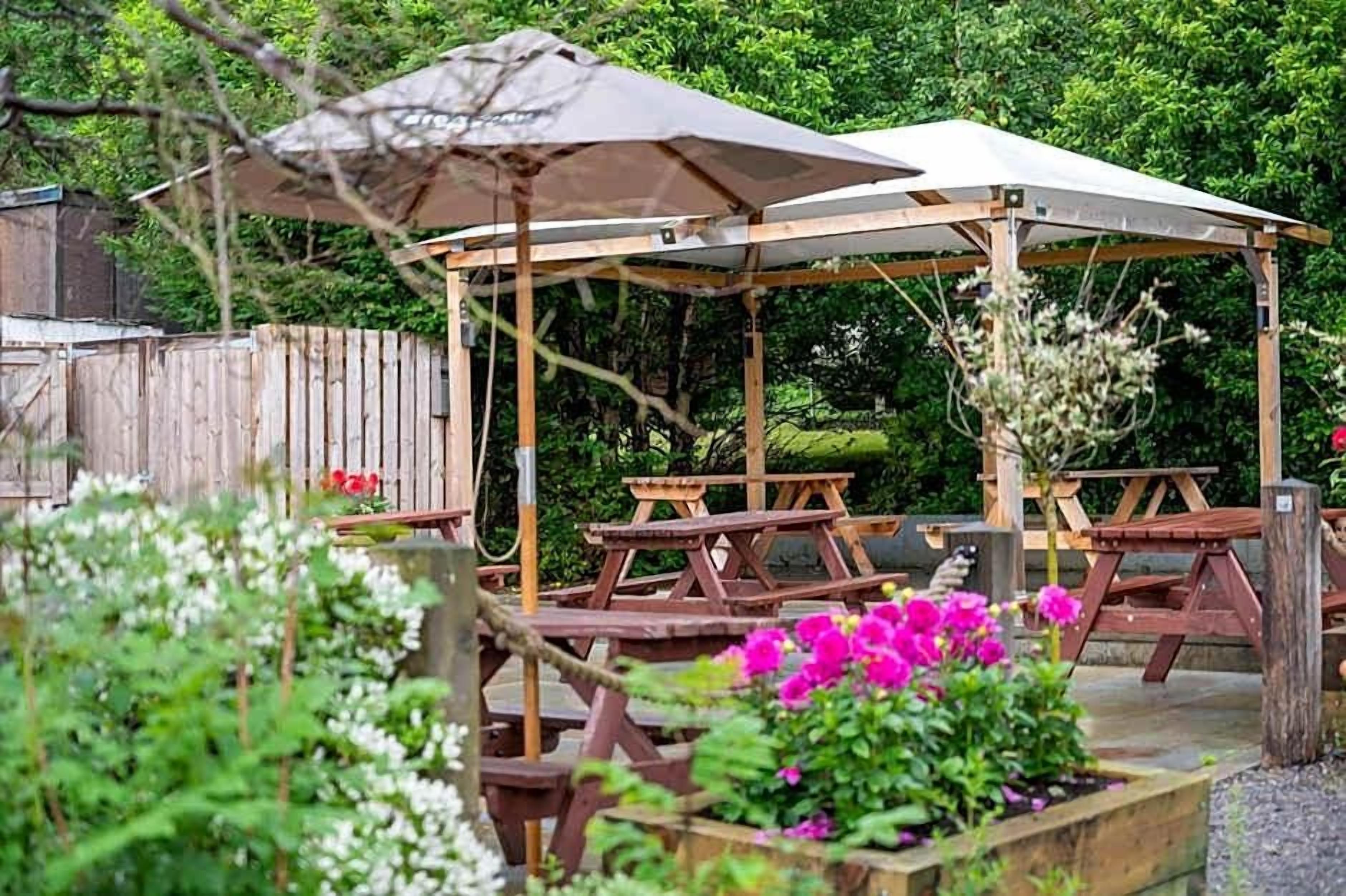 A wooden gazebo with a roof, surrounded by picnic tables and flower beds.