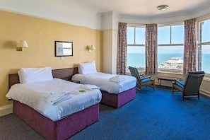 A hotel room with two beds, a small table, and a chair. There is a window with curtains and a view of the sea. - De Paris (Cromer)