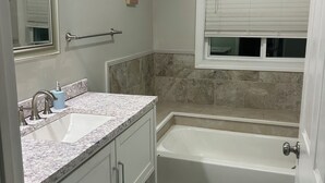 Un baño con lavamanos, espejo, tocador, bañera y ventana con persianas.