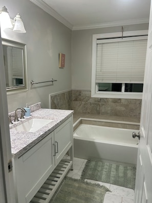 Un baño con lavamanos, espejo, tocador, bañera y ventana con persianas.
