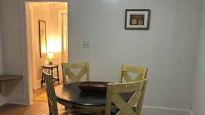 Un comedor con una mesa redonda, cuatro sillas de madera, una alfombra, un cuadro en la pared y una puerta que da a otra habitación.