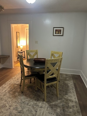 Un comedor con una mesa redonda, cuatro sillas de madera, una alfombra, un cuadro en la pared y una puerta que da a otra habitación.