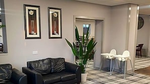 Um hall com piso xadrez, um sofá de couro preto, duas quadros, uma planta em vaso e uma pequena mesa com cadeiras.