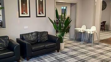 A lobby with a checkered floor, a black leather sofa, two framed pictures, a potted plant, and a small table with chairs.