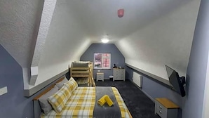Um quarto pequeno com telhado inclinado, uma cama, uma escrivaninha, uma cadeira e uma janela.