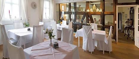 A dining area with multiple tables, white tablecloths, and chairs. There are windows with curtains, a chandelier, and a view of a Christmas tree.
