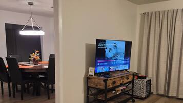 Una sala de estar con televisor sobre un mueble de madera, un comedor con mesa y sillas, y un pasillo que conduce a otra habitación.