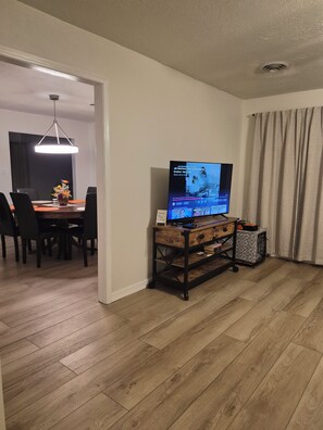 A living room with a television on a wooden stand, a dining area with a table and chairs, and a hallway leading to another room.
