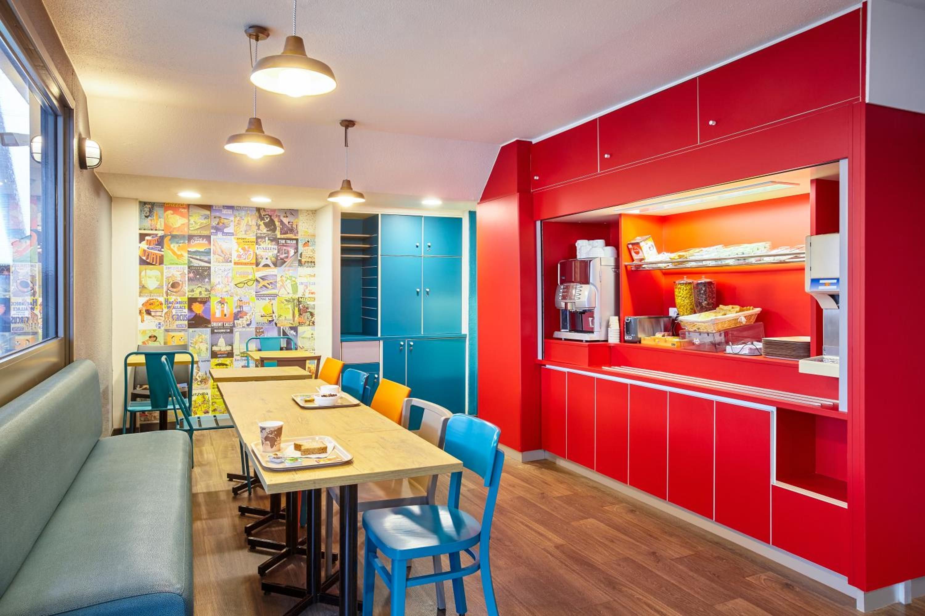 Un espace repas avec une longue table, des chaises bleues et un comptoir rouge sur lequel se trouve une machine à café.