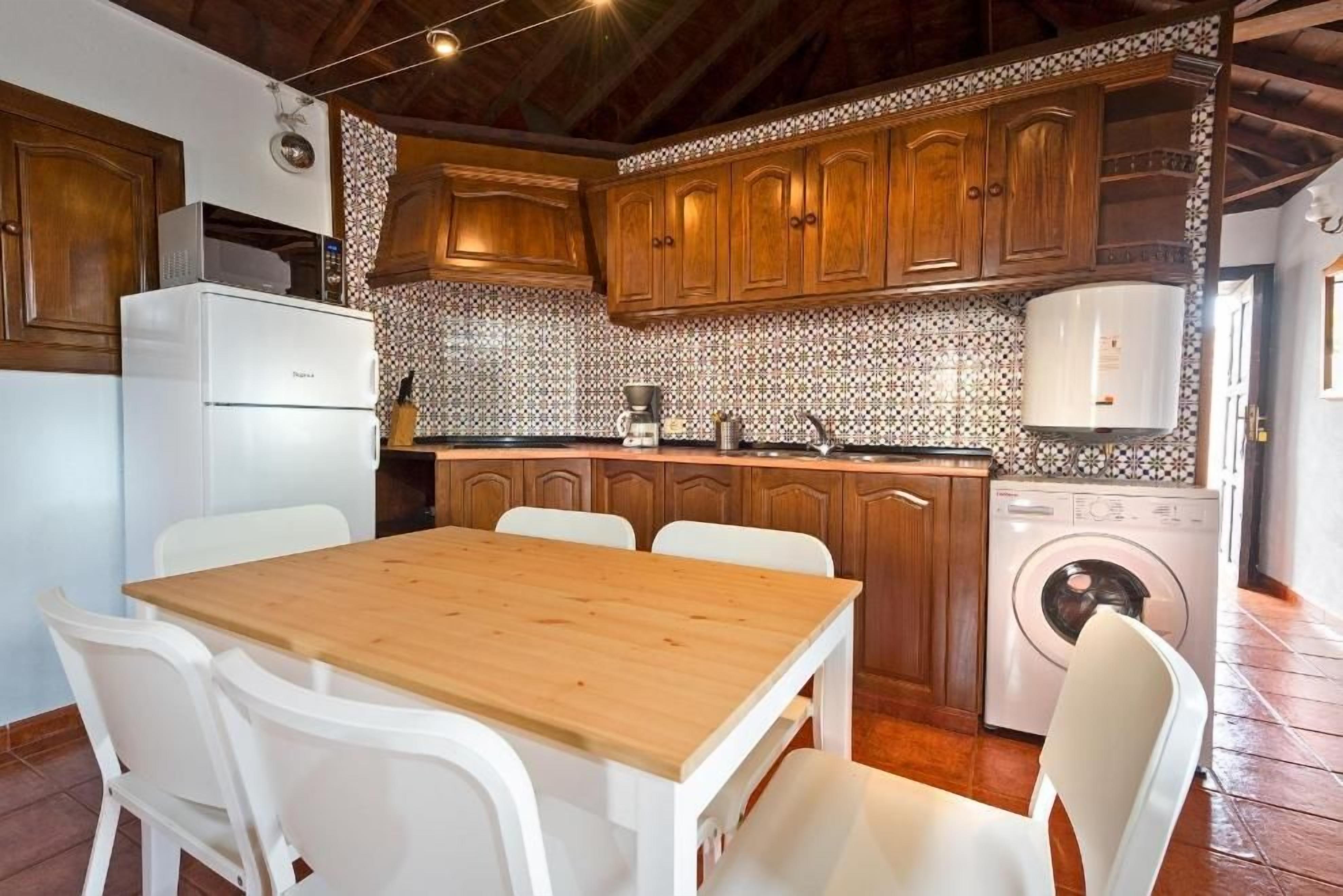 Una cocina con armarios de madera, un refrigerador blanco, una mesa de comedor de madera con sillas, una cafetera y una lavadora.