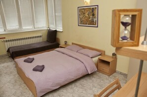 A bedroom with a bed, a bedside table, a nightstand, a radiator, a window with blinds, and a wall-mounted painting. - Hostel Mostel (Sofia)