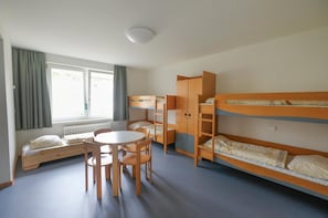 A dormitory room with bunk beds, a table, chairs, and a window with curtains.
