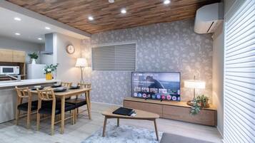 A modern living room with a wooden ceiling, a flat-screen TV, a dining area with a table and chairs, and a kitchen in the background.