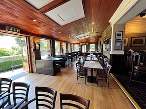 Restaurante com teto de madeira, mesas compridas e vista para o verde lá fora.