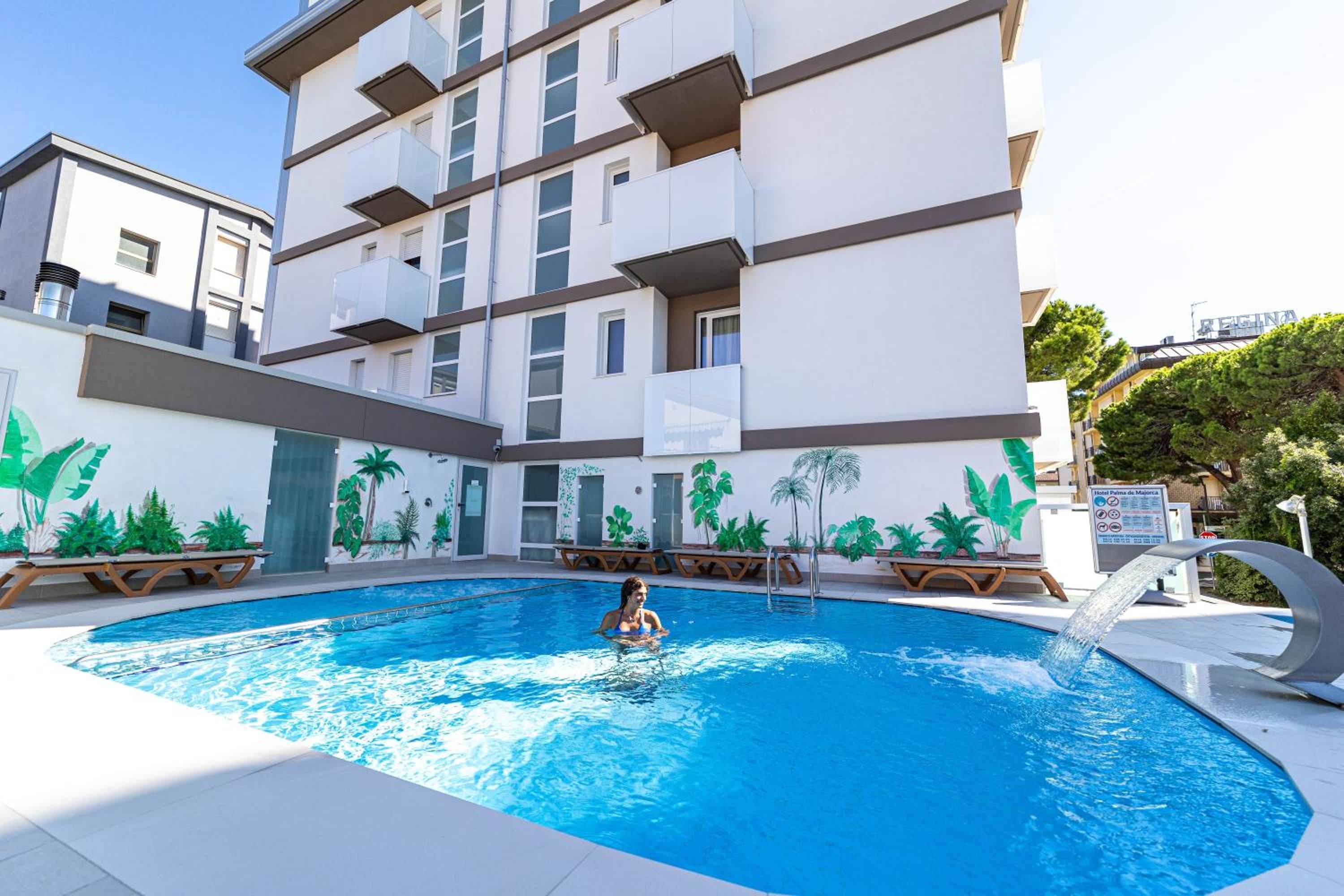 A swimming pool with a person in the water, surrounded by a building with balconies and a mural of tropical plants.