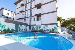 Una piscina con una persona a nuoto, circondata da un edificio con balconi e un murale di piante tropicali.
