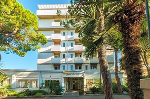 Un hotel a più piani con un ingresso centrale, circondato da palme e altra vegetazione.