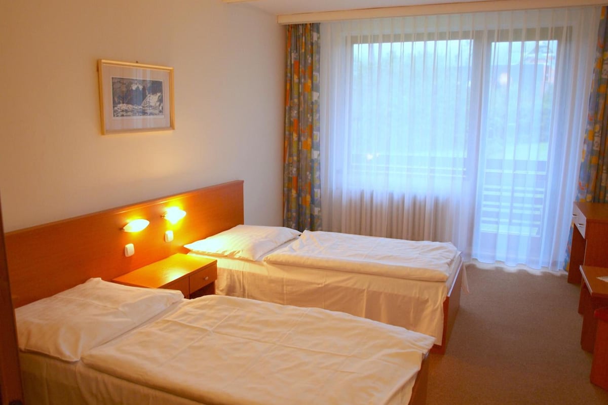Una camera d'albergo con due letti, un comodino con una lampada, un quadro appeso al muro e una finestra con tende leggere.