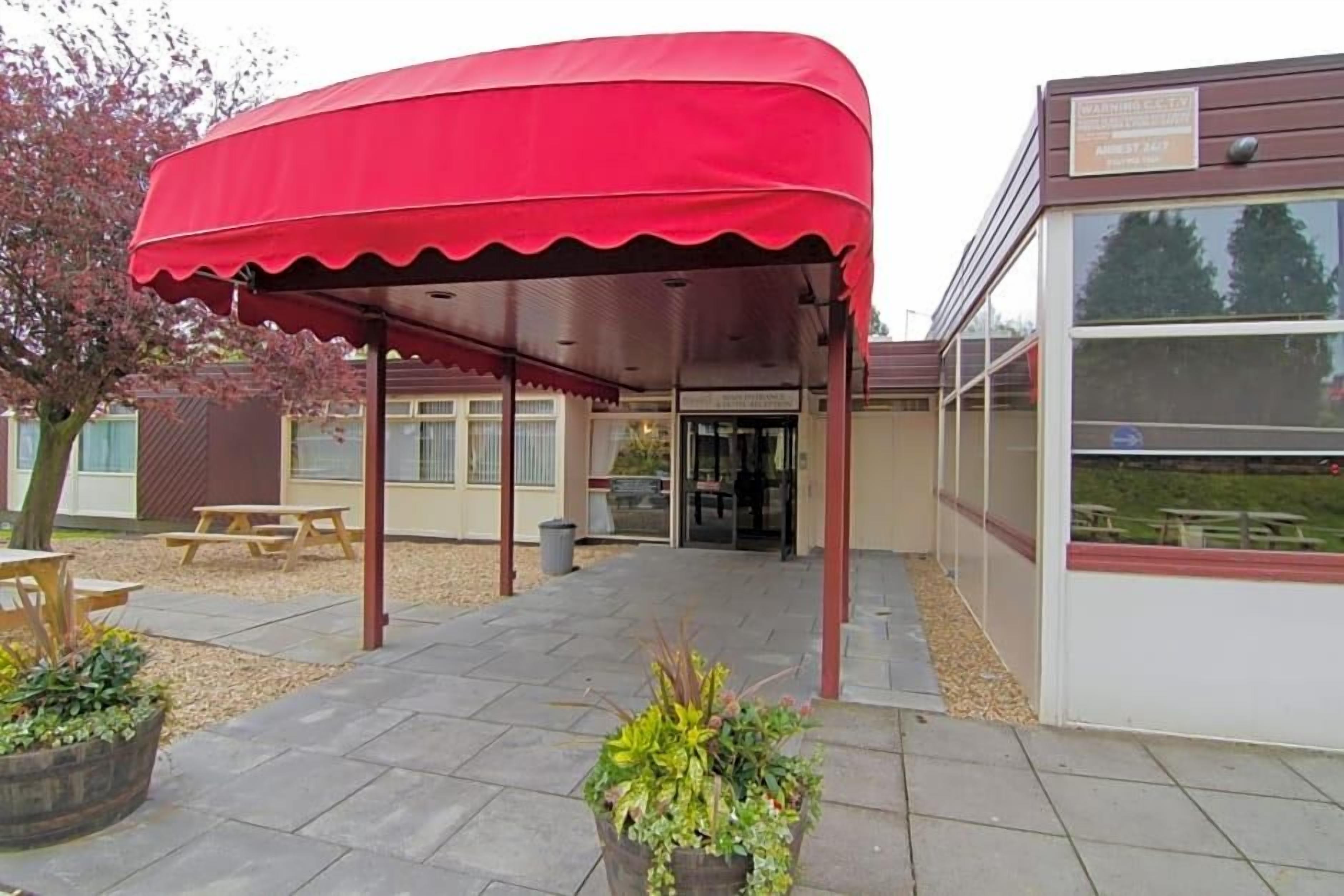 Een overdekt buitengebied met een rood dak, een picknicktafel en een klein gebouw met een bord.