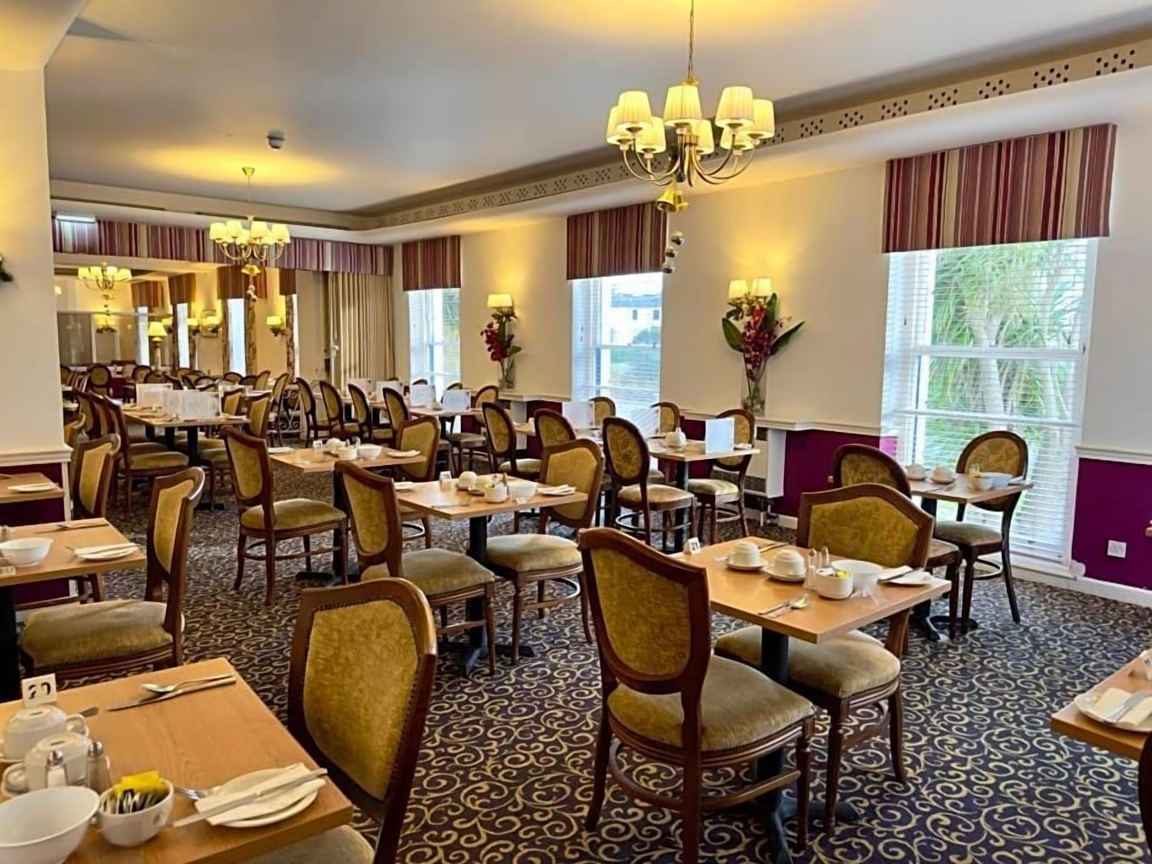 A hotel dining area with multiple tables and chairs, chandeliers, and large windows with curtains.