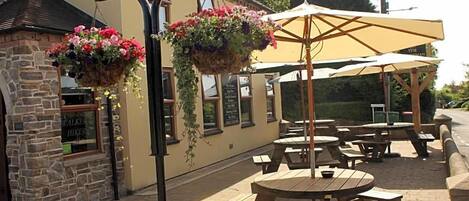 An outdoor seating area with tables and benches, a hanging flower basket, and a building with a stone facade.