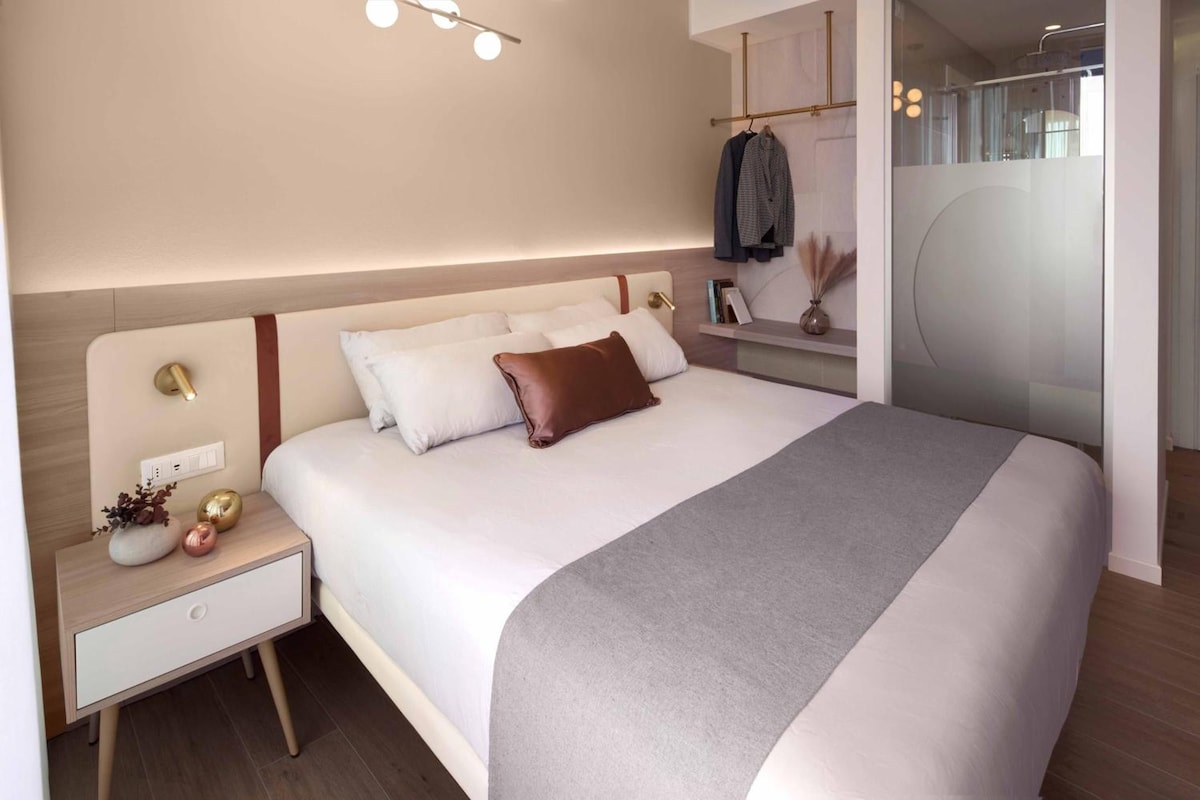 Un dormitorio moderno con una cama grande, mesitas de noche, un pequeño mueble auxiliar y un estante en la pared con libros y un florero.