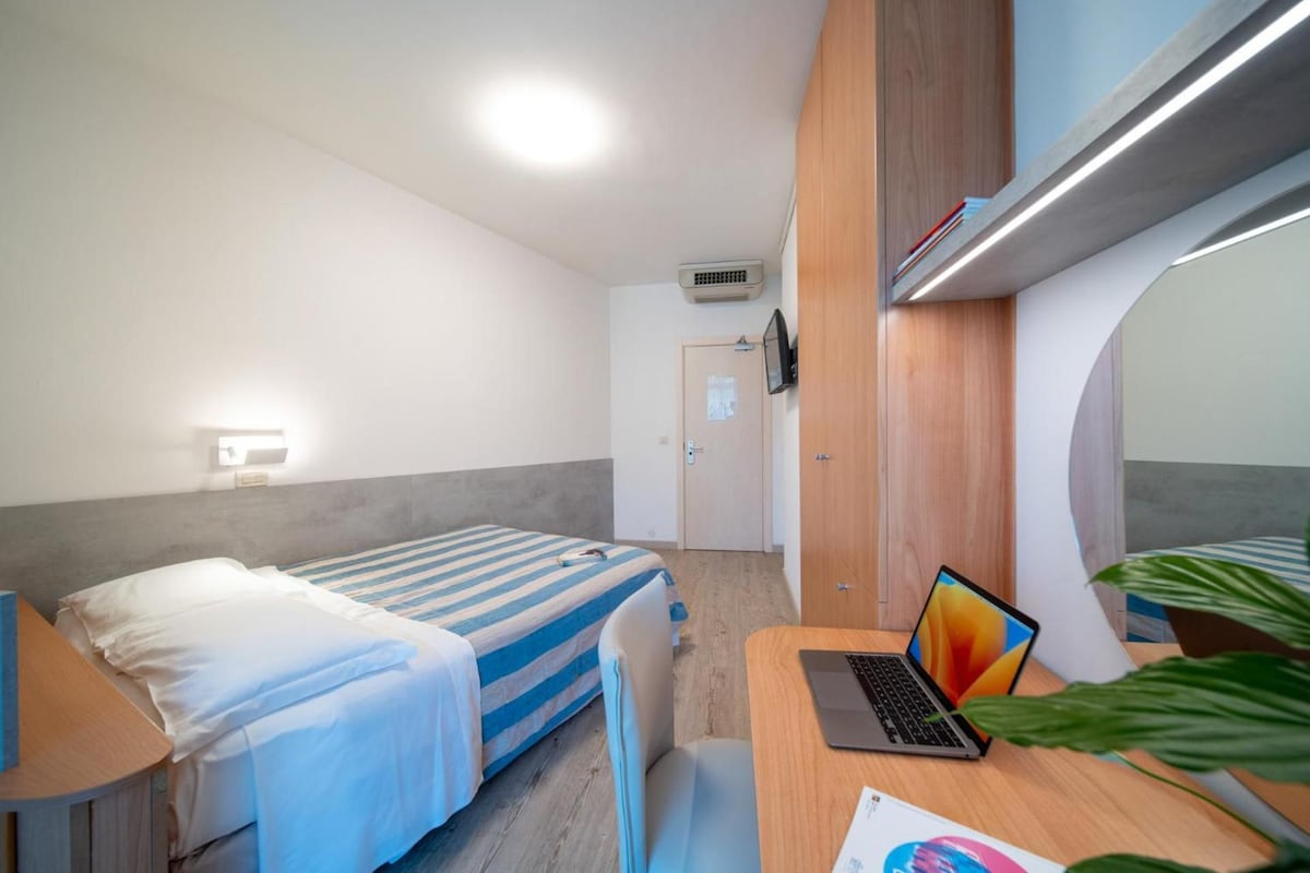 Una habitación de hotel con una cama, un escritorio con una computadora portátil, un espejo y una planta.