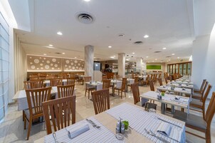 Un'ampia sala ristorante con diversi tavoli e sedie, una zona cucina sullo sfondo e un moderno design del soffitto.
