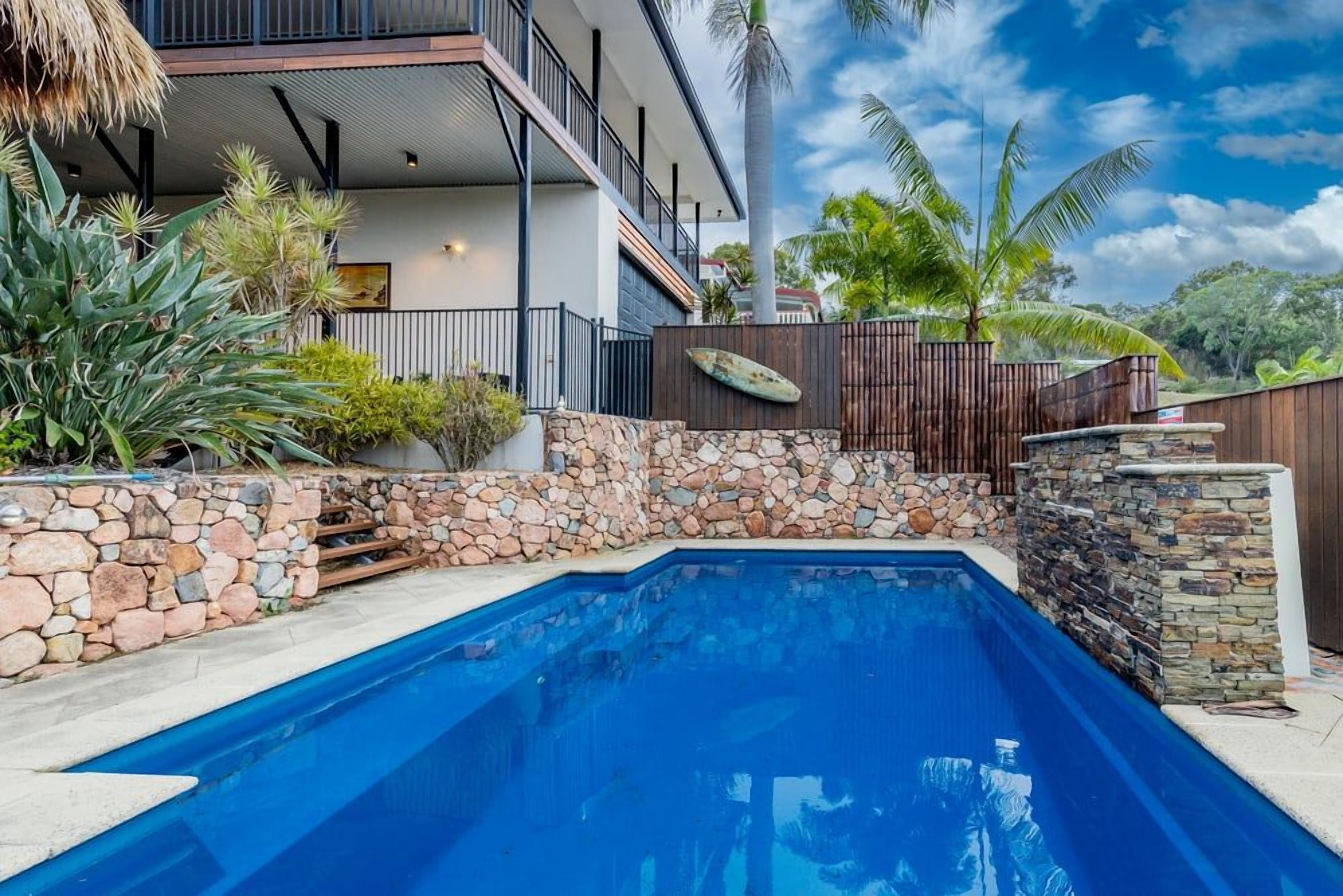 A swimming pool with a stone wall and a wooden fence, surrounded by tropical plants and a two-story building with balconies.