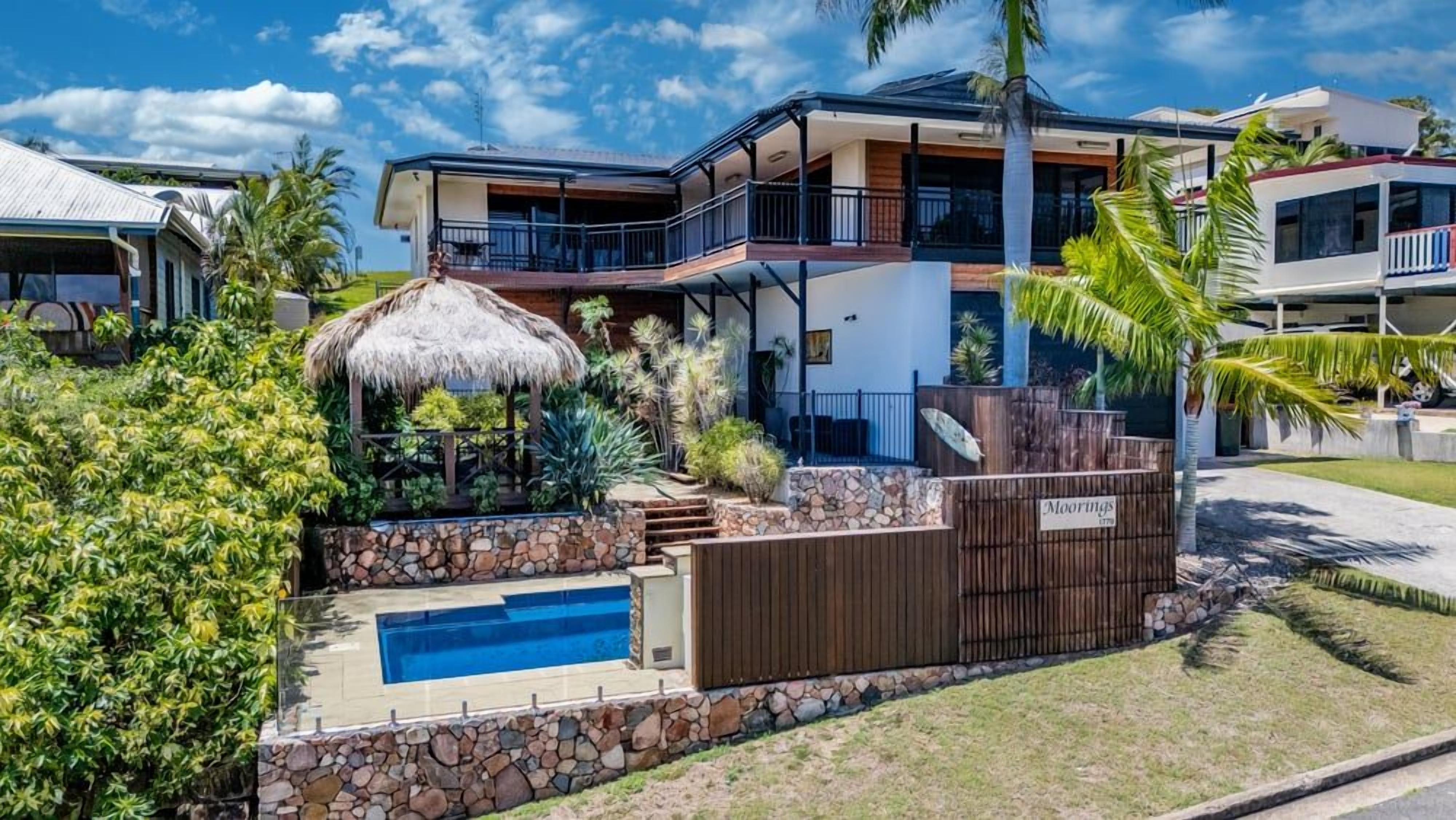 Ein modernes, zweistöckiges Haus mit einer Steinmauer, einem Swimmingpool und einem Gebäude mit einem Reetdach.