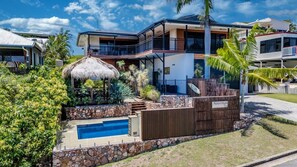 A modern two-story house with a stone retaining wall, a swimming pool, and a thatched-roof structure.