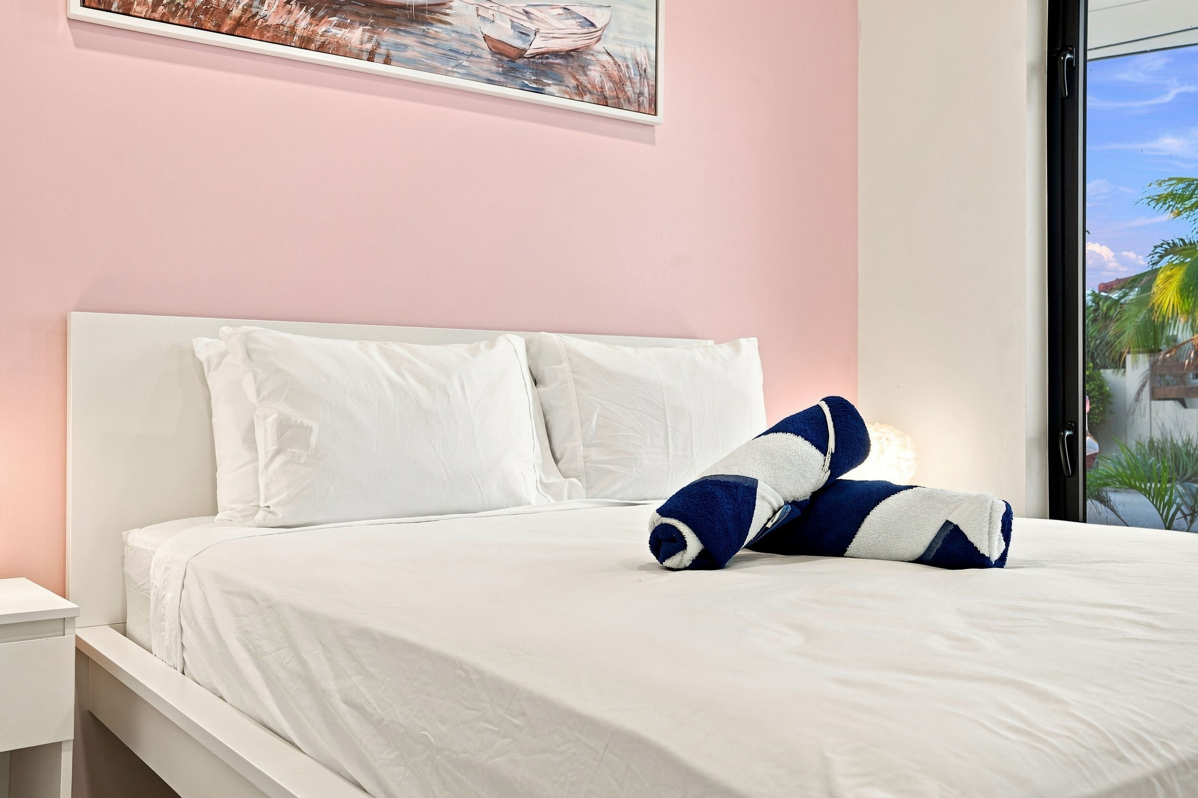 A bed with white bedding and two pillows, one white and one blue and white striped, in a room with a pink wall and a window.