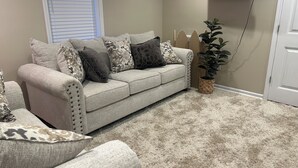 En stue med en beige sofa, mønstrede puder, en potteplante og et vægmonteret fladskærms-tv.