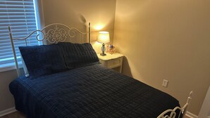 En seng med mørkt quiltet dyne, et hvidt metalhovedgærde, et natbord med lampe og et vindue med persienner.