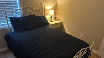 Una cama con colcha oscura acolchada, cabecero de metal blanco, mesita de noche con lámpara y ventana con persianas.