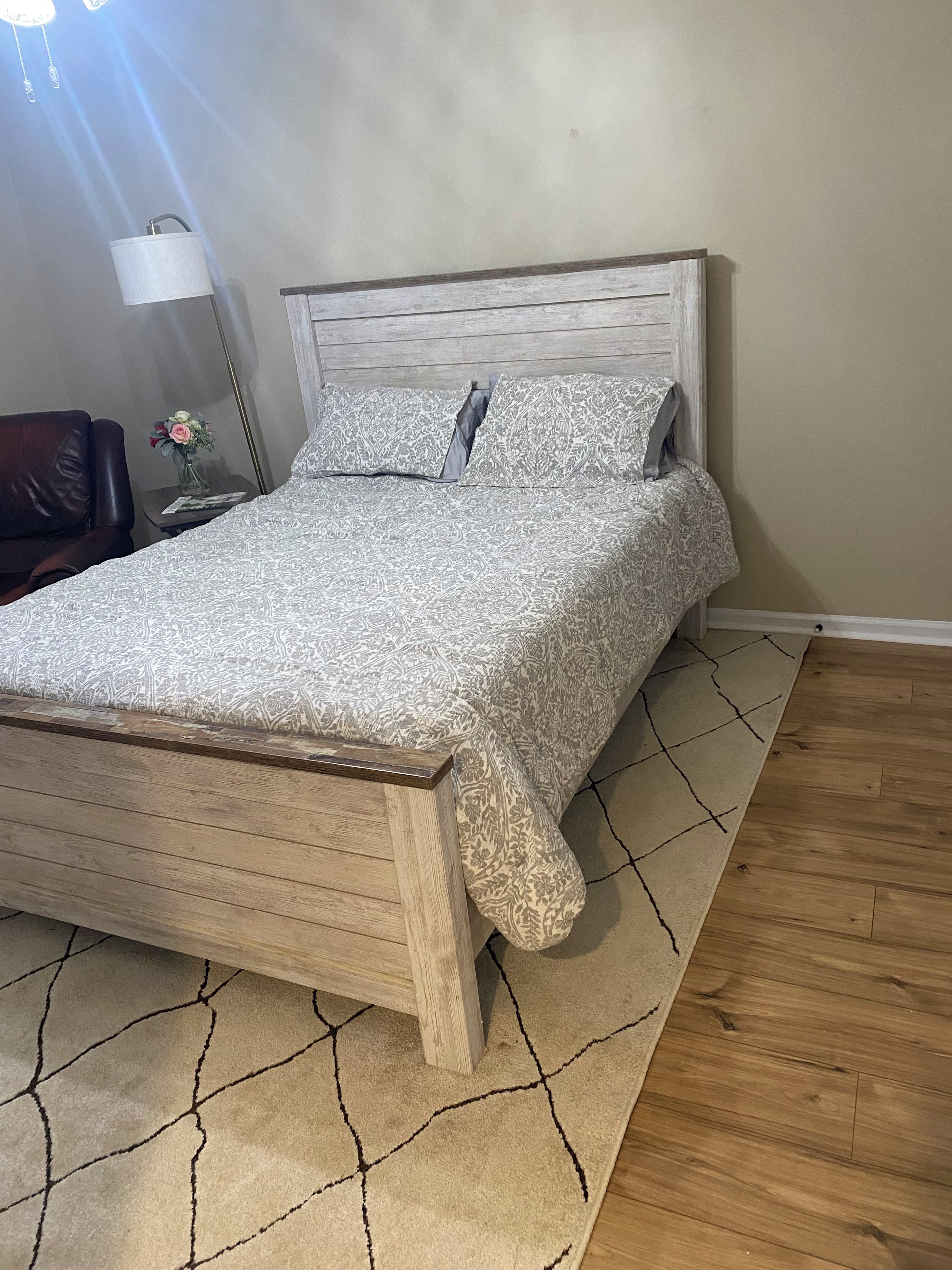 Una cama con colcha estampada, cabecera de madera y mesita de noche con lámpara y un florero.