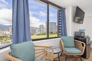 A room with two wicker chairs, a small table, a television mounted on the wall, and a view of buildings through a large window. - Waikiki Studio | Pool + Beach Gear | City Views (Honolulu)
