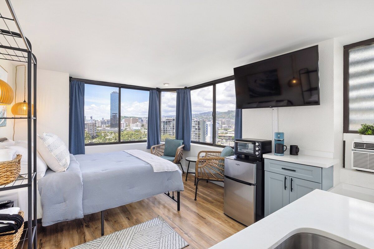 A compact room with a bed, kitchenette, and large windows offering a city view.