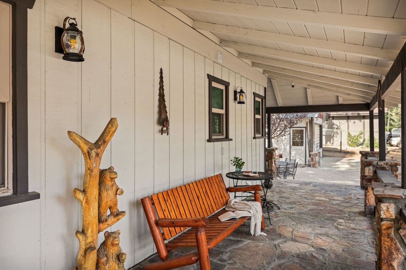 A covered porch with a wooden bench, stone flooring, and rustic decor including a bear sculpture and wall-mounted lanterns.