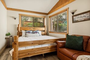 Ein Schlafzimmer mit einem hölzernen Bett, zwei Fenstern mit Jalousien, einem braunen Ledersofa und einem kleinen Nachttisch mit Lampe.